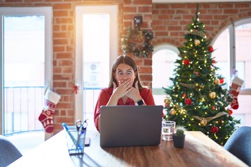 Beautiful woman sitting at the table working with laptop at home around christmas tree shocked covering mouth with hands for mistake. Secret concept.