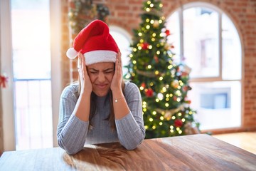 Young beautiful woman wearing santa claus hat at the table at home around christmas decoration suffering from headache desperate and stressed because pain and migraine. Hands on head.