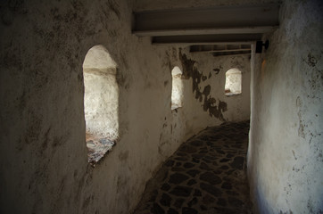 Gallery inside an old defensive structure. There are several loopholes on the left, a wall on the right, wooden beams under the ceiling. The gallery has a bend to the right. Background.