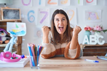 Young beautiful teacher woman wearing sweater and glasses sitting on desk at kindergarten celebrating surprised and amazed for success with arms raised and open eyes. Winner concept.