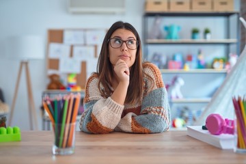 Young beautiful teacher woman wearing sweater and glasses sitting on desk at kindergarten with hand...