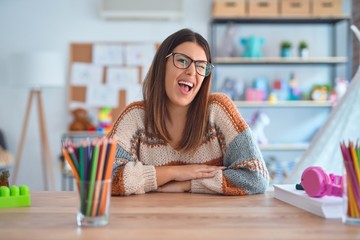 Young beautiful teacher woman wearing sweater and glasses sitting on desk at kindergarten winking...