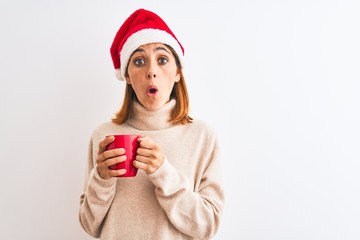 Beautiful redhead woman wearing christmas hat drinking a cup of coffee over isolated background scared in shock with a surprise face, afraid and excited with fear expression