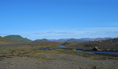 Icelandic lava desert landscape with bending river and panorama of Landmannalaugar colorful mountains and green hills. Fjallabak Nature Reserve, Iceland. Summer blue sky