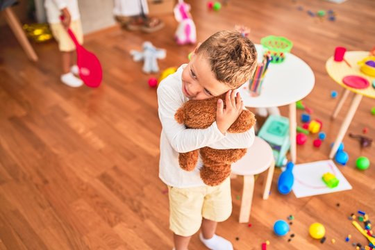 Beautiful blonde toddler hugging teddy bear standing around lots of toys at kindergarten