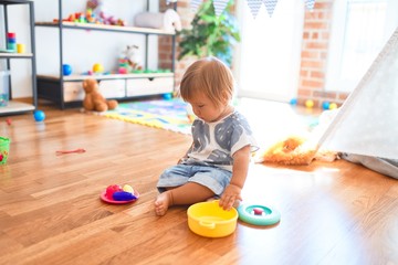Fototapeta premium Adorable toddler sitting on the floor playing meals using plastic cutlery and food around lots of toys at kindergarten