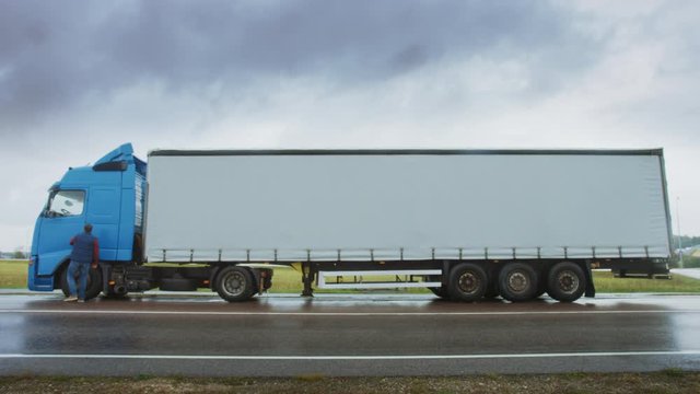 Truck Driver Crosses the Road in the Rural Area and Gets into His Blue Long Haul Semi-Truck with Cargo Trailer Attached. Logistics Company Moving Goods Across Countrie Continent. Side View Camera Shot