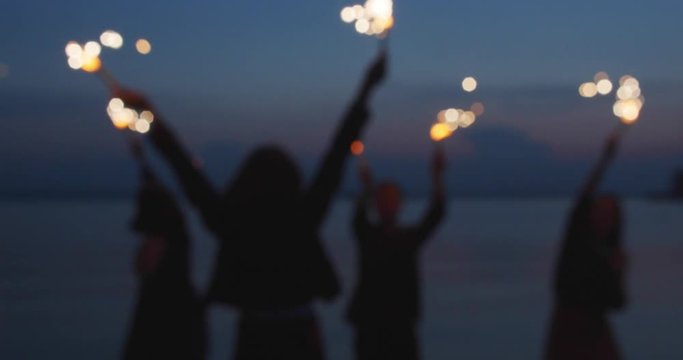 A company of friends celebrates with sparklers in their hands. Dancing, jumping, having fun. In the evening near the sea. Defocused, blurred picture