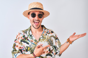 Fototapeta premium Young handsome man wearing Hawaiian shirt and summer hat over isolated background amazed and smiling to the camera while presenting with hand and pointing with finger.