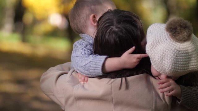 Back view of loving mother cuddling toddler son and daughter with down syndrome while spending leisure together in autumn park. Strong hugs of happy family with special child during freetime outdoor