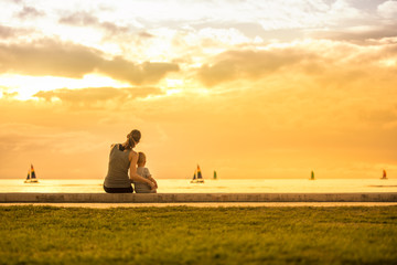 Mother and the child son sitting by the sea at sunset watching sailboats go by. 