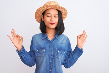 Obraz na płótnie Canvas Young beautiful chinese woman wearing denim shirt and hat over isolated white background relax and smiling with eyes closed doing meditation gesture with fingers. Yoga concept.