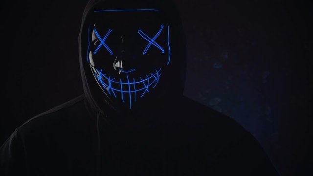 Man with lighting neon glow mask in hood raises his head and scream in camera on black background. Halloween and horror concept