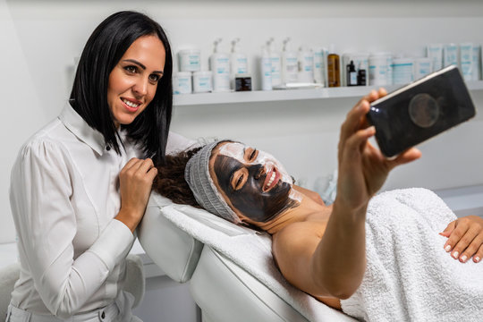 Beautiful woman receiving facial mask with rejuvenating effects in spa beauty salon and taking selfie photo with her doctor, beautician  expert.