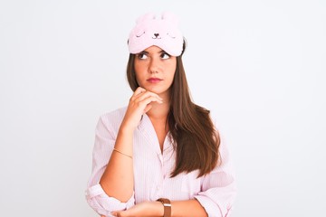 Young beautiful girl wearing pajama and sleep mask standing over isolated white background with hand on chin thinking about question, pensive expression. Smiling with thoughtful face. Doubt concept.