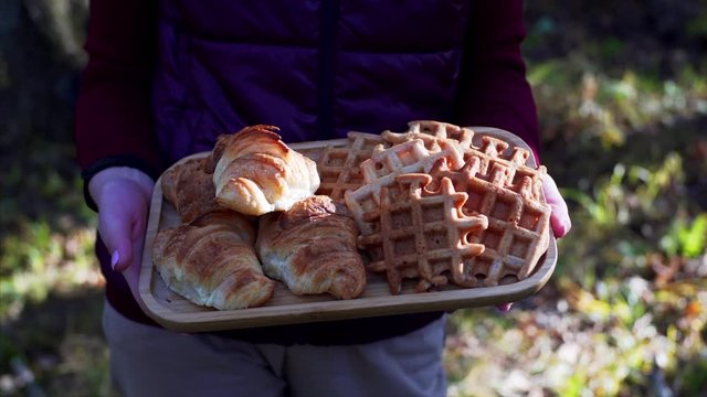 Unidentified woman is holding tray with fresh belgian waffles and croissants cooked for picnic in autumn forest