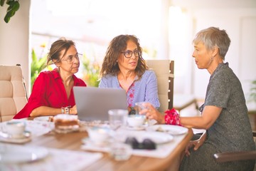 Meeting of middle age women having lunch and drinking coffee. Mature friends smiling happy using laptop at home on a sunny day