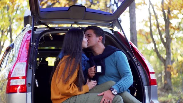 Young couple is sitting in car trunk with hot tea in metal cups and chatting and kissing. Autumn forest