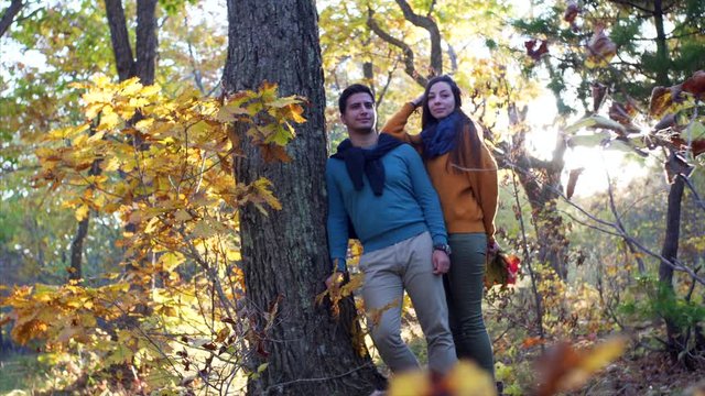 Attractive man and woman in love are posing near big tree in sunny autumn forest