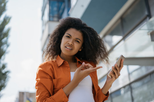 Emotional African American woman pointing finger on cellphone screen. Young excited hipster girl using mobile application for online shopping with cash back, low prices. Female ordering food online