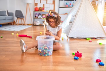 Beautiful toddler wearing glasses and unicorn diadem sitting on the floor playing with building blocks at kindergarten