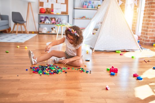 Beautiful toddler wearing glasses and unicorn diadem sitting on the floor playing with building blocks at kindergarten