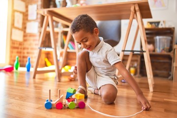 Fototapeta premium Beautiful african american toddler playing with wooden blocks train toy around lots of toys at kindergarten