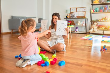 Beautiful psycologist and blond toddler girl sitting on the floor doing therapy using emoji emotions at consulting room