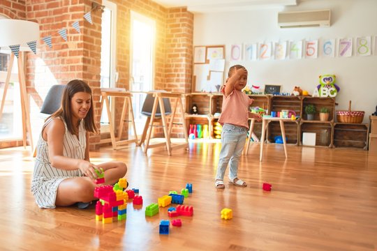 Beautiful teacher and blond toddler girl building tower using plastic blocks at kindergarten