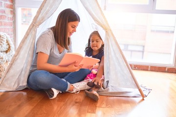 Beautiful teacher reading book to toddler girl sitting on the floor inside tipi at kindergarten