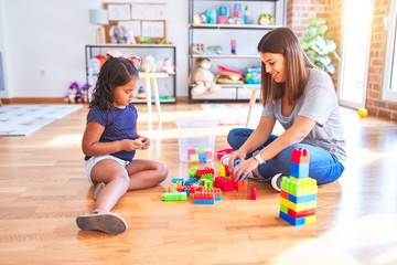 Fototapeta premium Beautiful teacher and toddler girl playing with construction blocks bulding tower at kindergarten
