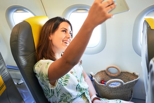 Young traveller woman sitting inside plane at the airport with sky view from the window taking a selfie with smartphone