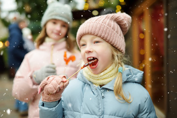 Two adorable sisters having rooster-shaped lollipops on traditional Christmas fair in Riga, Latvia. Children enjoying sweets, candies and gingerbread on Xmas market.