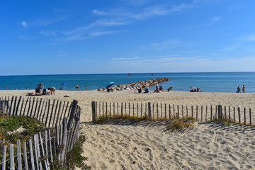 Fototapeta premium Holiday makers enjoy the sun on a sandy beach in south of France. No lifeguard. Calm sea and pier at the background. 