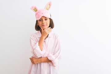 Beautiful child girl wearing sleep mask and pajama standing over isolated white background looking confident at the camera with smile with crossed arms and hand raised on chin. Thinking positive.