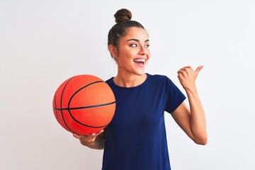 Young beautiful sportswoman holding basketball ball over isolated white background pointing and showing with thumb up to the side with happy face smiling