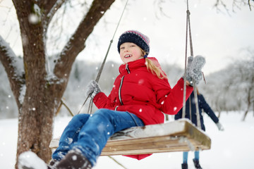 Two adorable young girls having fun on a swing together in beautiful winter park. Cute sisters...