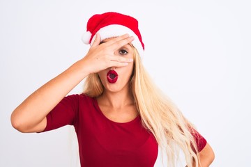 Young beautiful woman wearing Christmas Santa hat over isolated white background peeking in shock covering face and eyes with hand, looking through fingers with embarrassed expression.