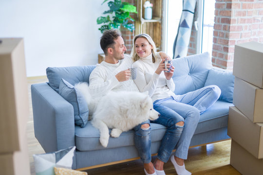 Young beautiful couple with dog sitting on the sofa drinking coffee at new home around cardboard boxes