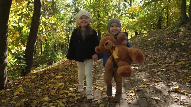Happy toddler boy with teddy bear and his older sister with down syndrome walking holding hands during family leisure in autumn park. Cheerful little kids enjoying freetime in nature on sunny day