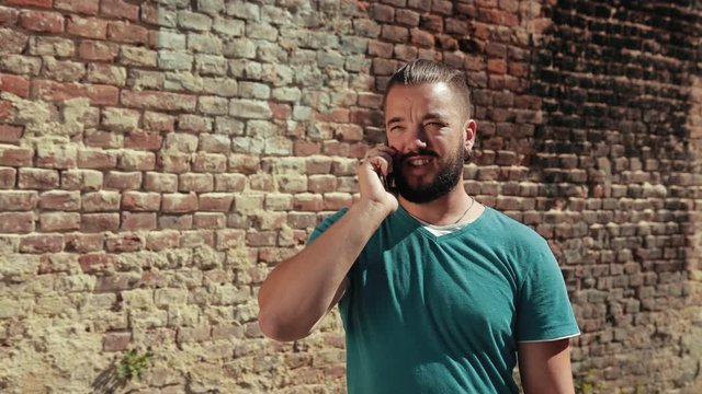 Young man with dark hair and beard, blue t-shirt holding smartphone near brick wall talking on the phone, happily and with smile, sunny weather