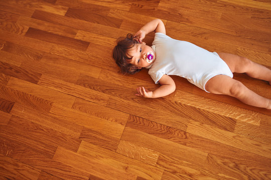 Beautiful toddler child girl wearing white bodysuit lying down on the floor using pacifier