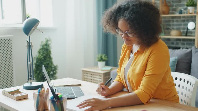 Cheerful teenager attractive young African American lady is using laptop at home typing then taking notes in notebook. People, gadgets and homework concept.