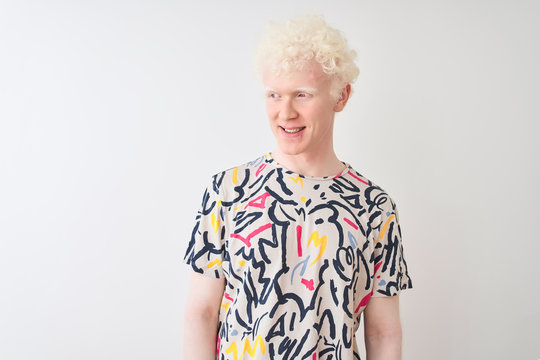 Young albino blond man wearing colorful t-shirt standing over isolated white background looking away to side with smile on face, natural expression. Laughing confident.