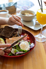 Woman eating breakfast with knife and fork in a cafe: avocado and nuts, rye toasts, vegetables, fried bacon, chicken and poached egg