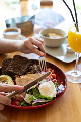 Woman eating breakfast with knife and fork in a cafe: avocado and nuts, rye toasts, vegetables, fried bacon, chicken and poached egg