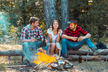 Naklejka na ściany i meble Friendship and leisure concept. Friends hikers watching fire together on camp. Weekend. Good day for spring picnic in nature. Beautiful young friends enjoying picnic in a park.