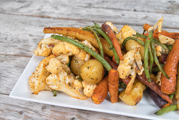 Roasted vegetables on a white platter on wooden table. Carrots, potatoes, herbs, cauliflower and green beans. Overhead view