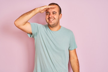 Obraz premium Young man wearing blue casual t-shirt standing over isolated pink background very happy and smiling looking far away with hand over head. Searching concept.