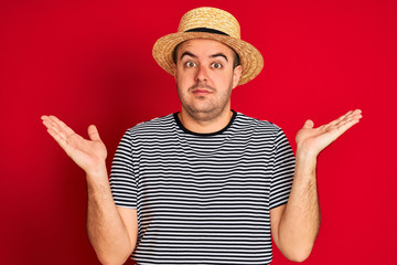 Young man wearing striped navy t-shirt and hat standing over isolated red background clueless and confused expression with arms and hands raised. Doubt concept.